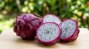 Un fruit du dragon entier et trois moitiés coupées, révélant la chair blanche parsemée de graines noires, sur une planche en bois, avec un arrière-plan flou de verdure.